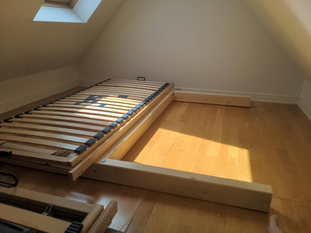 the built bed frame, an “H” made from wooden beams. one of two slotted frames is put on the left half, the other side is empty. you can see a screw with some washers at each end, to keep the frames in place.
