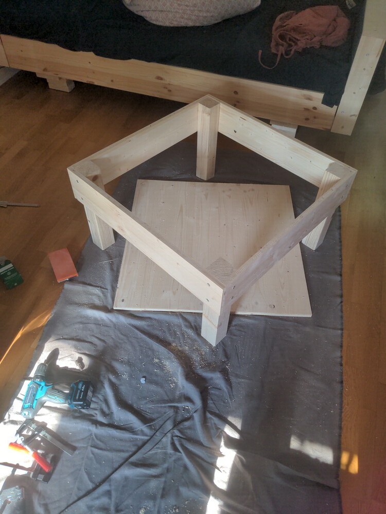 the built frame for the stool. the seating area is still missing, its lies below. everything on a big grey sheet of fabric. next to it a drill, screw clamps, saaaaaawdust. in the background you can see the sofa from the other blogpost.